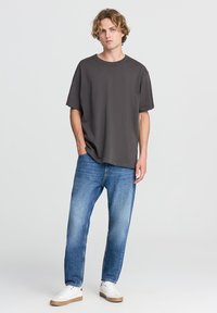 T-shirt oversize grigio scuro realizzata in tessuto morbido, abbinata a jeans a gamba dritta blu chiaro e sneakers bianche con accenti beige.