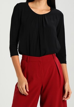 Femme portant un chemisier noir à manches trois-quarts rentré dans un pantalon rouge taille haute à jambes larges sur un fond uni.