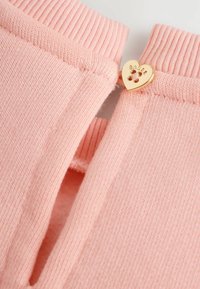 Maglione rosa chiaro a maglia con polsini a coste e un bottone dorato a forma di cuore come dettaglio su uno spacco. La texture è morbida e liscia.