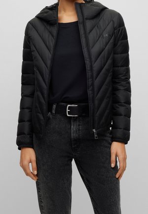 Persona con chaqueta acolchada negra con capucha, camiseta negra, cinturón negro y vaqueros negros desgastados, frente a un fondo claro y liso.