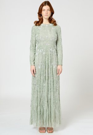 Langes grünes Kleid mit langen Ärmeln, verziert mit silberner Stickerei und fließenden Falten, kombiniert mit hellfarbigen Sandalen.