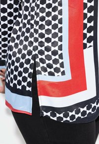 Blusa de patrones en blanco y negro con formas geométricas, acentos en rojo y azul claro, abertura lateral y textura de tela suave.