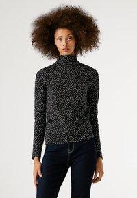 Egy nő voluminózus, göndör hajjal, fekete, hosszú ujjú garbóban fehér pöttyökkel és sötétkék farmernadrágban áll egy fehér háttér előtt.