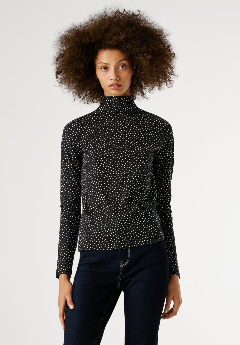 Egy nő voluminózus, göndör hajjal, fekete, hosszú ujjú garbóban fehér pöttyökkel és sötétkék farmernadrágban áll egy fehér háttér előtt.