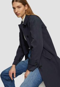 Marineblauwe trenchcoat met een getailleerde snit, grote knopen en zijzakken, gedragen over een witte top en gecombineerd met lichtblauwe jeans.