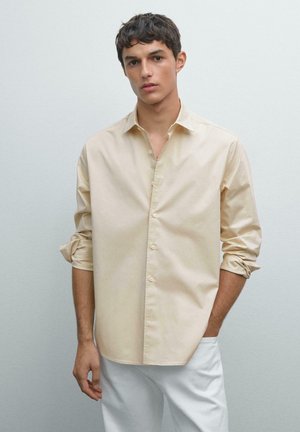 Camicia button-up in cotone crema con colletto classico, maniche lunghe arrotolate fino ai gomiti e una vestibilità rilassata. Abbinata a pantaloni bianchi.