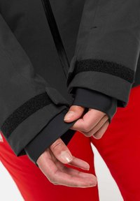 Des mains ajustant le poignet d'une veste noire avec des bandes Velcro sur une manche intérieure noire, un pantalon rouge visible en arrière-plan.