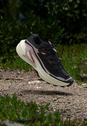 Chaussure de trail Salomon noire et rose avec une semelle épaisse suspendue au-dessus d'un chemin de terre avec de l'herbe et du feuillage en arrière-plan.