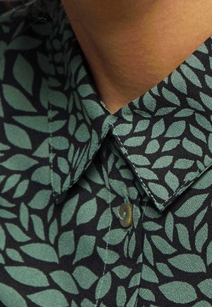 Tmavá košile se zelenými listovými vzory, zapnutý límec, nošená osobou se středně hnědou pokožkou viditelnou v oblasti krku.