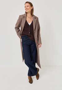 Lungo cappotto di lana marrone con risvolti, indossato sopra un cardigan viola scuro e una maglietta bianca, abbinato a jeans blu scuro e stivali alla caviglia con stampa leopardo.