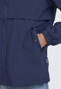Navy windbreaker jacket with a zip front, elastic cuffs, side pockets with snaps, and a smooth, lightweight fabric.