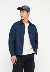 Giacca di jeans blu scuro con chiusura a bottoni, taschino sul petto e cuciture a contrasto. Indossata sopra una camicia bianca e abbinata a pantaloni neri e un cappellino blu.