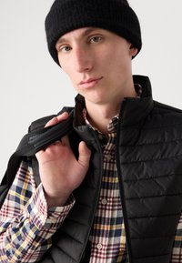 Colete puffy preto com costura horizontal, combinado com uma camisa xadrez multicolorida com botões brancos e colarinho redondo. Gorro de malha preto usado.