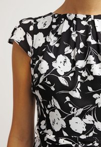 Svart klänning med vitt blommönster, plisserad halsringning och korta ärmar. Tyget har en mjuk textur och en figurnära silhuett.
