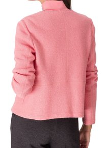 Femme portant une veste rose pâle texturée à manches longues sur un pantalon gris foncé, vue de dos avec les bras légèrement pliés.