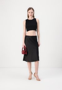 Svart crop top kombinert med en flytende svart midi-skjørt, holdt med en rød håndveske. Modellen har på seg nude ankelrem-sandaler, mot en ensfarget bakgrunn.