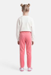 Παιδί με ξανθά μαλλιά σε δύο κότσους, που φοράει άσπρο μπλουζάκι με μακριά μανίκια, ροζ και άσπρο παντελόνι και ροζ αθλητικά παπούτσια, στέκεται γυρισμένο προς τα πίσω.