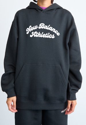 Osoba nosící černou mikinu New Balance a odpovídající černé tepláky s bílým logem, stojící před obyčejným světlým pozadím.