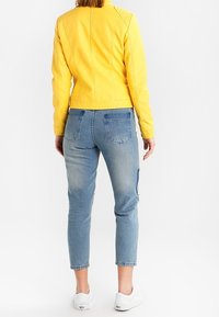 Giacca di pelle gialla con dettagli testurizzati sulle spalle, abbinata a jeans di denim blu chiaro e sneakers bianche. Vista da dietro.