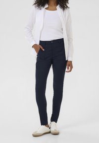 Femme portant des baskets blanches, un pantalon tapered bleu marine avec poches, un débardeur blanc et une chemise blanche déboutonnée, se tenant contre un fond uni.