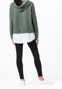 Zielona przycięta bluza z kapturem, biała warstwowa koszula, czarne legginsy i białe trampki. Gładkie tekstury, casualowy design, luźny krój i detal kaptura.