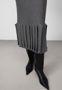 Grijze geplooide rok van gestructureerde stof, met gelaagde plooien aan de zoom, gecombineerd met hoge zwarte puntige laarzen.