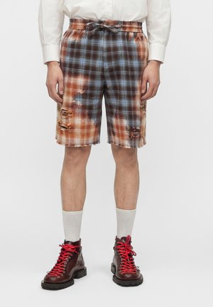 Καρό σορτς με φθαρμένες τρύπες και κορδόνι, φορεμένα με λευκές κάλτσες και σκούρα καφέ μπότες με κόκκινα κορδόνια.