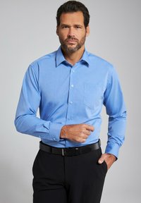Camisa de manga larga de color azul claro con cuello, cierre frontal de botones y un bolsillo en el pecho. Tejido suave con una textura sutil, combinada con pantalones negros.