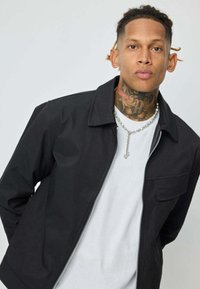 Un homme avec des tatouages sur le cou et des cheveux tressés porte une veste noire sur une chemise blanche et un collier en chaîne argenté, posant devant un fond gris clair.