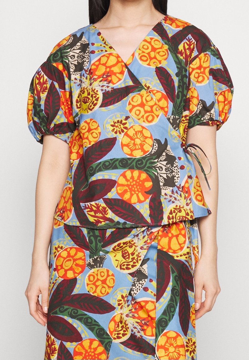 Haut portefeuille coloré avec un fond bleu, des motifs floraux vibrants orange et rouge, des manches bouffantes, et un détail noué à la taille.