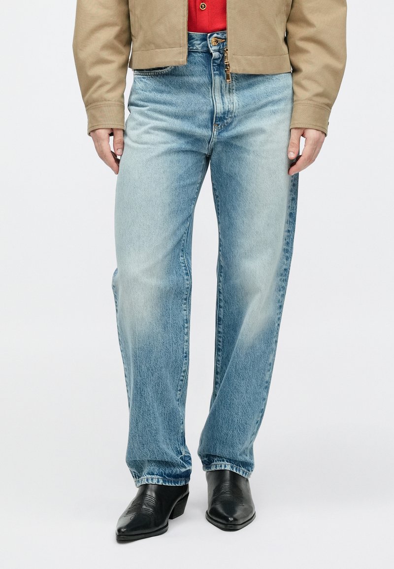 Man draagt lichtblauwe rechte jeans, zwarte leren puntige laarzen, beige jas en rode shirt, staand tegen een witte achtergrond.