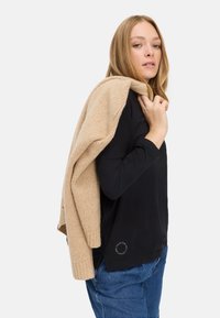 Schwarzes Langarmshirt mit einem kreisförmigen Akzent am Saum, kombiniert mit einem leicht beigen gestrickten Pullover, der über die Schulter gelegt ist. Blaue Jeans.
