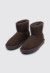 Botines de gamuza marrón con un forro interior suave, punta redondeada y suela de goma texturizada para mayor agarre. Diseño minimalista, sin herrajes visibles.