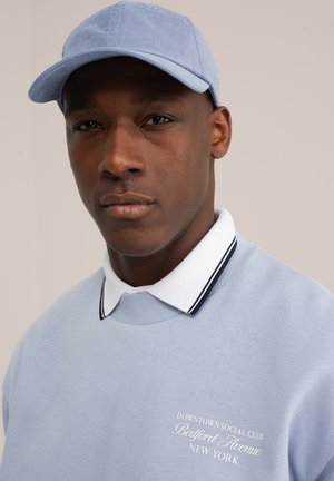 Man draagt een lichtblauwe pet en bijpassende sweatshirt met wit boordhemd, op de sweatshirt staat de tekst "Downtown Social Club Bedford Avenue New York."