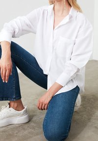 Camicia bianca con colletto e maniche lunghe, taschino sul petto; abbinata a jeans blu e sneakers bianche. Tessuto morbido, vestibilità comoda.