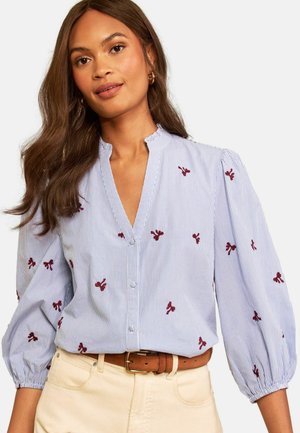 Femme aux longs cheveux bruns portant un chemisier rayé bleu et blanc avec une broderie florale rouge et un pantalon beige avec une ceinture marron.