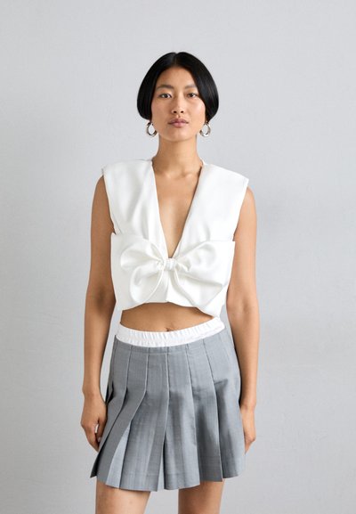Top court blanc avec un décolleté en V profond et un grand nœud à l'avant ; associé à une jupe mini plissée grise avec une taille élastique blanche.