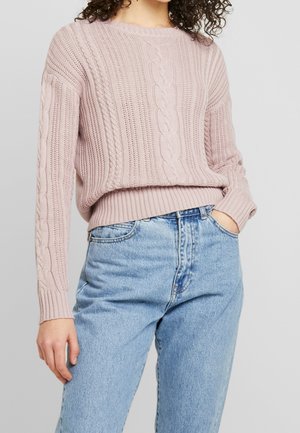 Personne portant un pull en maille torsadée rose clair et un jean taille haute bleu clair, une main dans la poche, sur un fond uni gris clair.