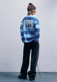 Modrá a námornícka pásikavá košeľa s dlhým rukávom a bielym nápisom "PRIATELIA NEKLAMU," spárovaná s čiernymi širokými nohavicami a hnedou pletenou čiapkou.