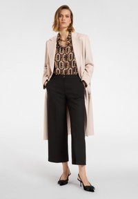 Jasnoróżowy długi płaszcz na wzorzystej brązowo-żółtej bluzce, w połączeniu z czarnymi szerokimi spodniami i czarnymi szpilkami z kokardkami.