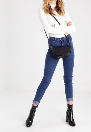 Černá kožená kabelka přes rameno s pleteným vzorem, kulatého tvaru a nastavitelným popruhem, doplněná modrými úzkými džínami a černými botami na podpatku.