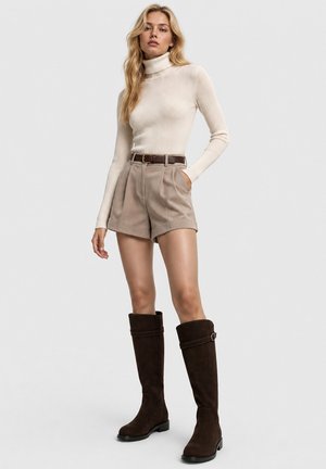 Mujer con un suéter de cuello alto color crema, shorts beige de cintura alta con un cinturón marrón, y botas largas de color marrón oscuro hasta la rodilla, de pie sobre un fondo liso.