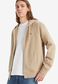 Beige zip-up hoodie met een trekkoord capuchon, grote voorzakken en een rood Levi's logo op de linker borst, gecombineerd met een wit t-shirt.