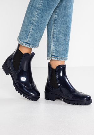 Kiiltävät tummansiniset Chelsea-saappaat, joissa on brogue-yksityiskohtia, ja joita käytetään käännettyjen vaaleansinisien farkkujen kanssa valkoisella lattialla.