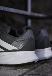 Une paire de chaussures de sport noires et grises avec des semelles blanches, posées sur un pavé en pierre à l'extérieur.