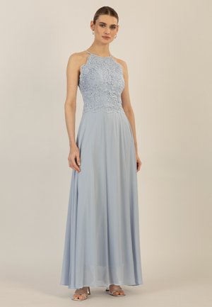 Frau steht und trägt ein ärmelloses, hellblaues Abendkleid mit floraler Spitze am Oberteil und fließendem Chiffonrock, kombiniert mit silbernen Sandalen.