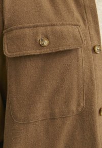 Brown wool shirt featuring a flap pocket with a button closure, textured fabric, and contrasting buttons. Simple, functional design.