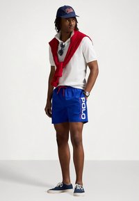Lässiges Outfit mit einem weißen Poloshirt, einem roten Pullover um den Hals gebunden, blauen Shorts mit "Polo"-Logo und marineblauen Sneakern.