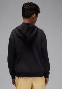 Black hoodie with a soft texture and a large hood, worn with loose beige trousers. Designed for a relaxed fit, featuring ribbed cuffs.