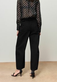 Schwarze Jeanshose mit weitem Bein und zwei Gesäßtaschen. Kombiniert mit einer schwarzen, transparenten Bluse mit Polka-Dots und schwarzen Sandaletten mit hohen Absätzen.
